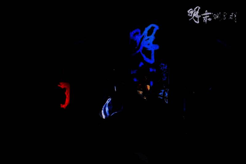 Coser扮演《明末：渊虚之羽》女主角无常，身穿黑色劲装，佩戴半面面具，站在展台前摆出战斗姿态，周围观众围观拍照