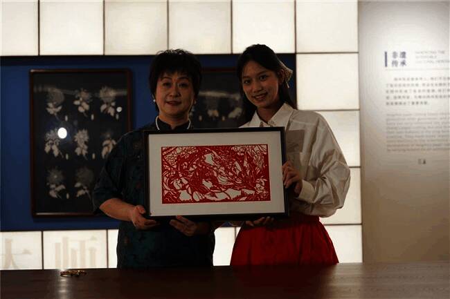 非遗传承人张震梅与博主苏久承在展览现场共同展示剪纸创作过程，周围观众围观拍摄