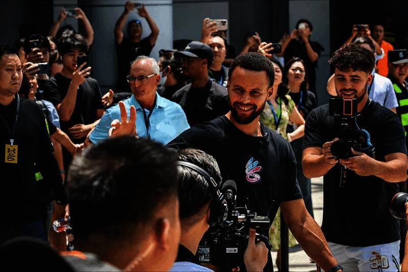 库里在展台前与工作人员交流，面带微笑，背景是巨大的安德玛品牌展墙