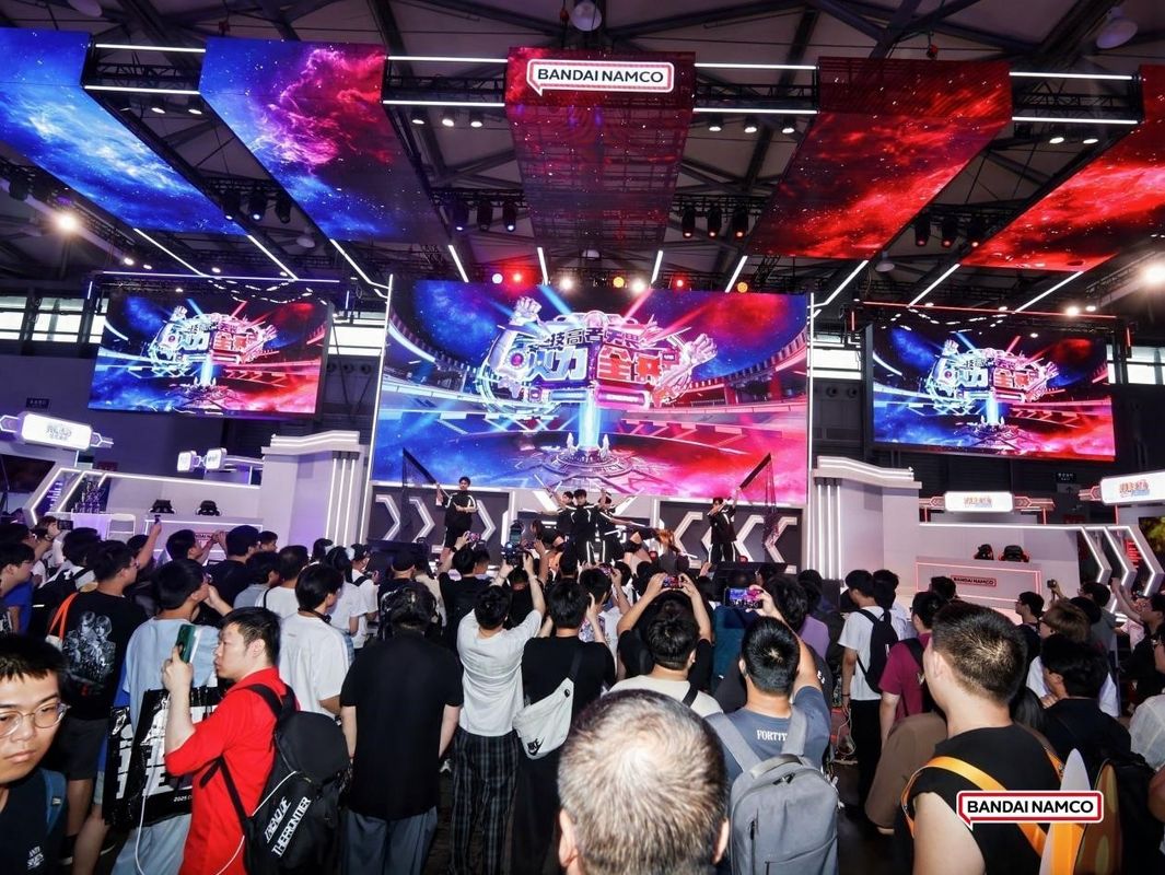 万代南梦宫展台全景，色彩绚丽，人潮涌动，舞台LED屏播放着游戏宣传片