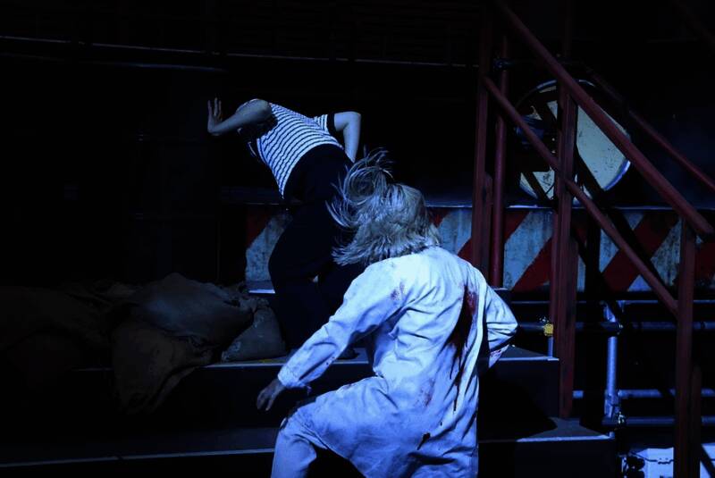 舞台灯光聚焦于吉尔持枪瞄准的特写，眼神坚定，背景为闪烁的警报红光