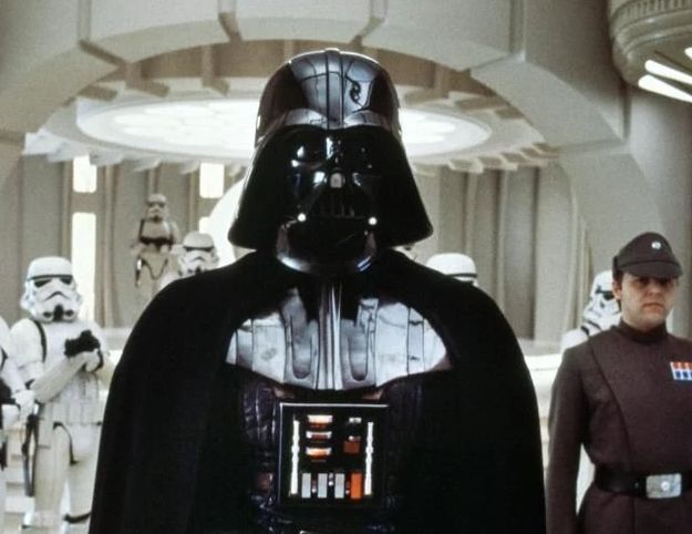 《星球大战》中达斯·维德手持标志性格斗姿态的光剑，剑身散发红色光芒，背景为暗色调星舰内部场景