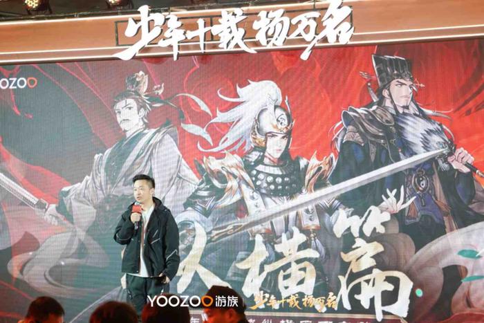 戴奇站在大屏幕前介绍《少年三国志3》概念图，屏幕上展示新风格武将原画与战斗界面