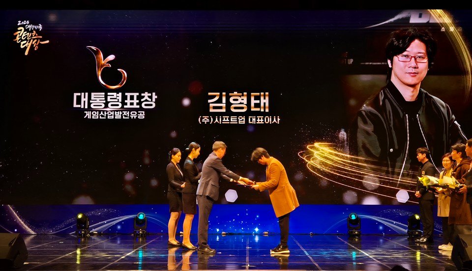 金亨泰在工作室内与团队成员讨论项目细节，背景屏幕上显示游戏角色设计图